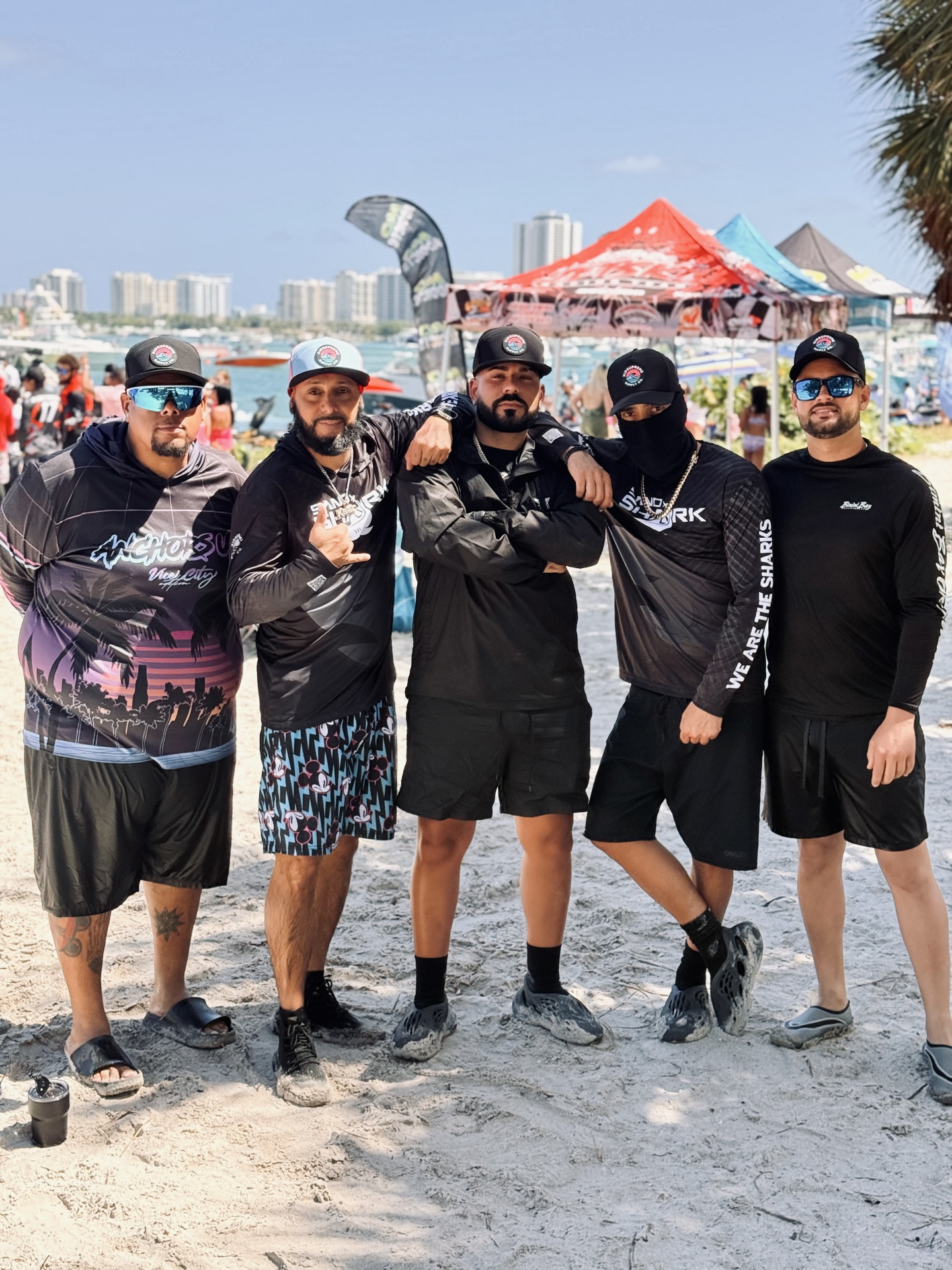 Group of five men in dark athletic wear posing on a sunny beach with tents and a city skyline in the background.
