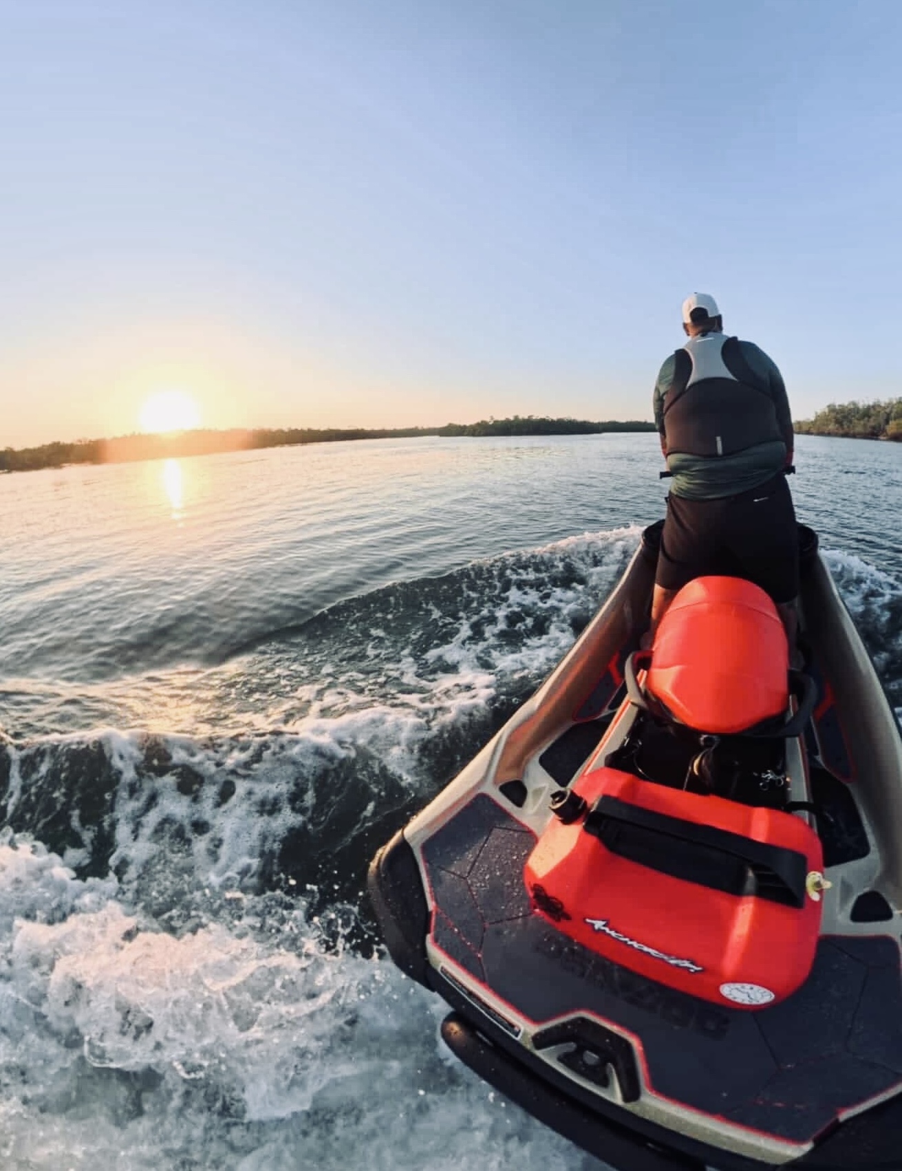 Person riding a red and black jet ski on calm water during sunset, wearing a life jacket and helmet, viewed from behind.