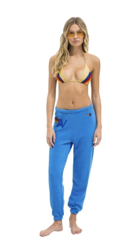 Model wearing a rainbow-striped bikini top and bright blue jogger sweatpants, with yellow sunglasses, standing against a white background.