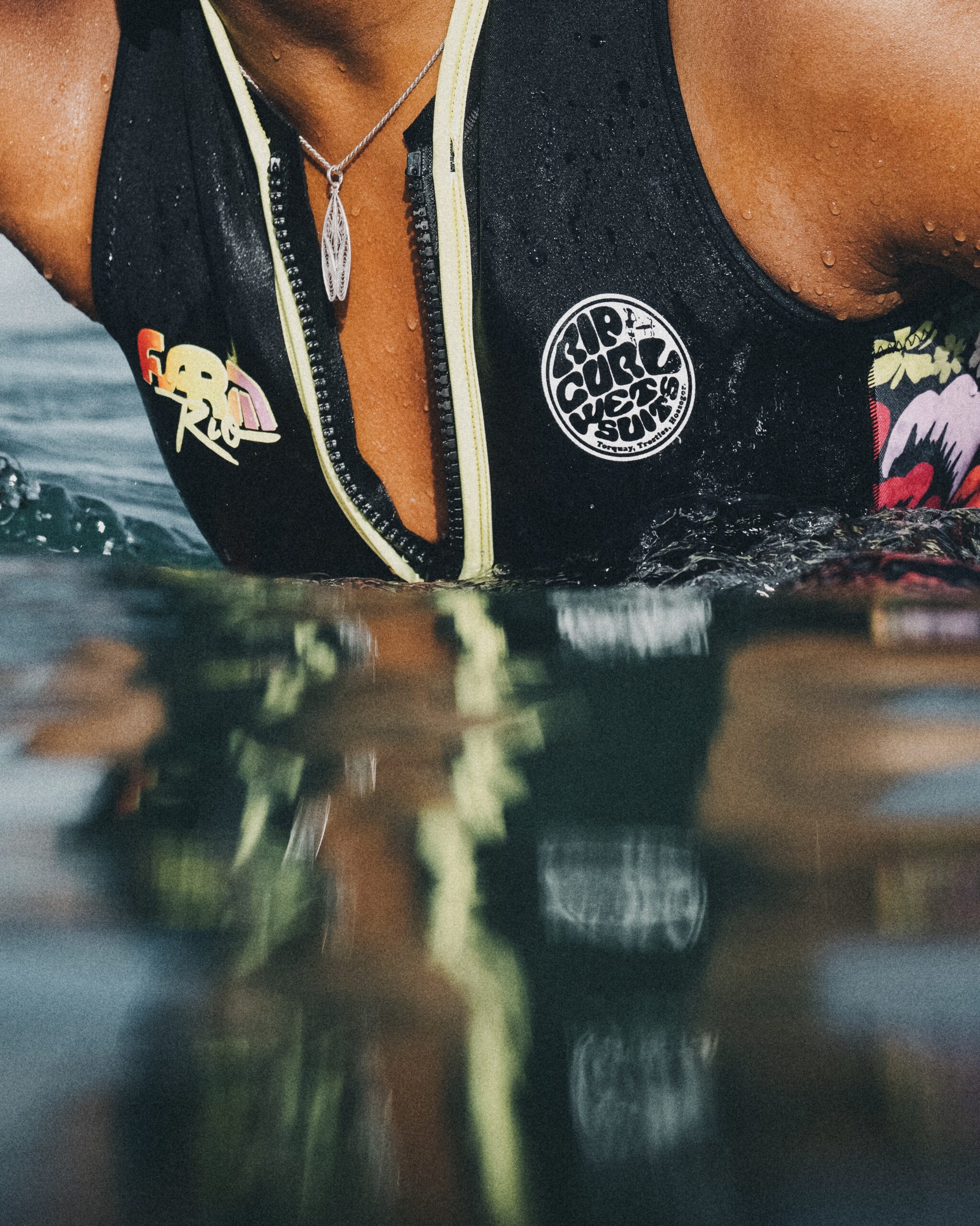 Close-up of a person wearing a dark wetsuit with a pale zipper, water droplets on their chest, and a pendant necklace. Ocean waves are visible in the background.