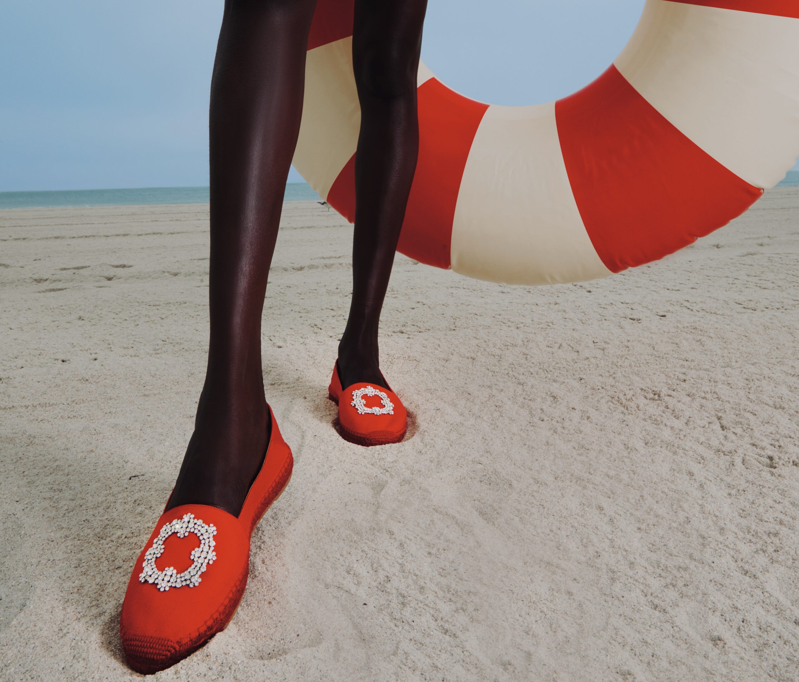 Close-up of a person’s legs in red shoes walking on a sandy beach with a red-and-white inflatable ring in the background.
