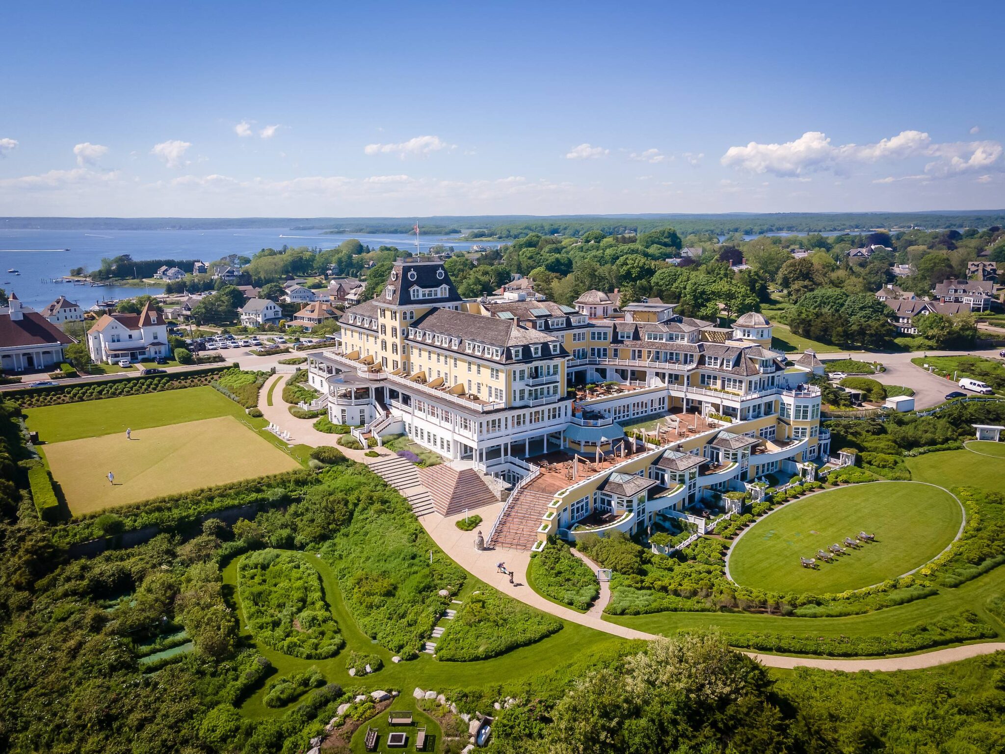 Aerial view of a seaside resort town with a large yellow hotel, manicured lawns, and a blue bay in the background under a clear sky.