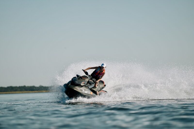 Person riding a jet ski across the water, creating a spray behind them, wearing a life jacket and helmet.