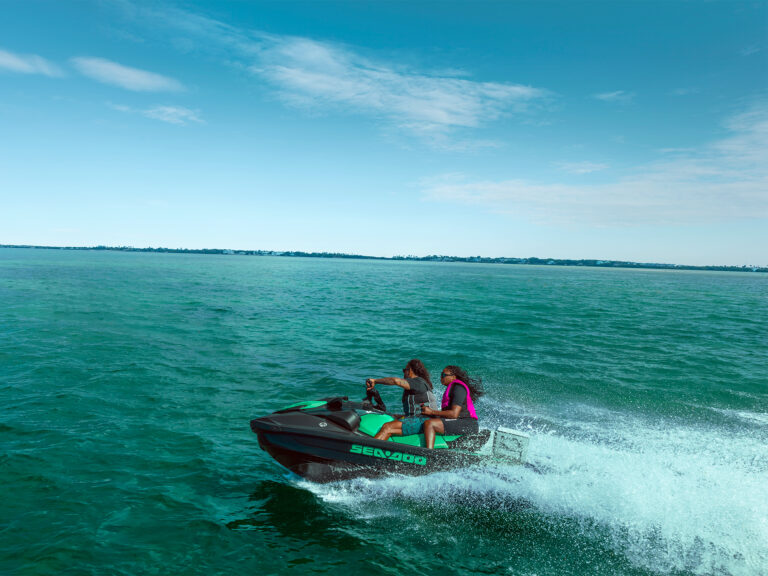 Two people ride a green Sea-Doo jet ski at speed on turquoise water, with a distant shoreline and blue sky visible on the horizon.