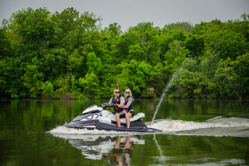 Two people riding a Yamaha jet ski on a calm lake with dense green trees in the background and a splash of water.