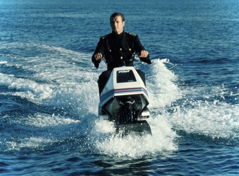 Man in a dark uniform riding a white jet ski across blue ocean water, creating a wake and spray.