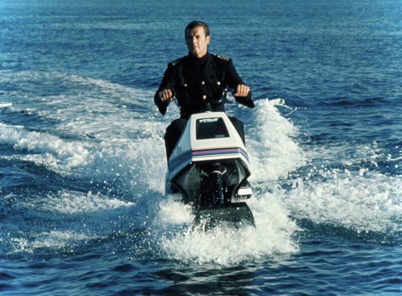 Man in a dark uniform riding a white jet ski across blue ocean water, creating a wake and spray.
