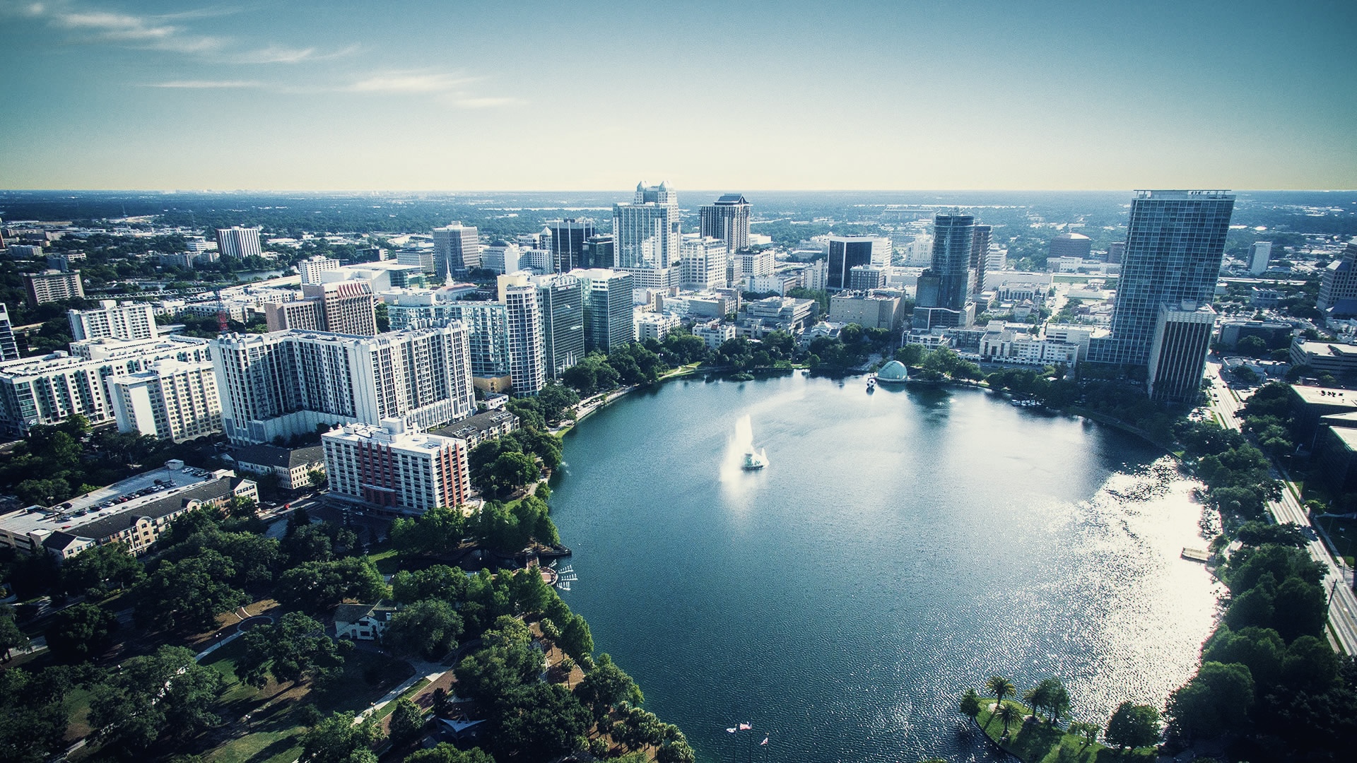 Aerial view of a downtown city surrounding a circular lake with a fountain and green park spaces.