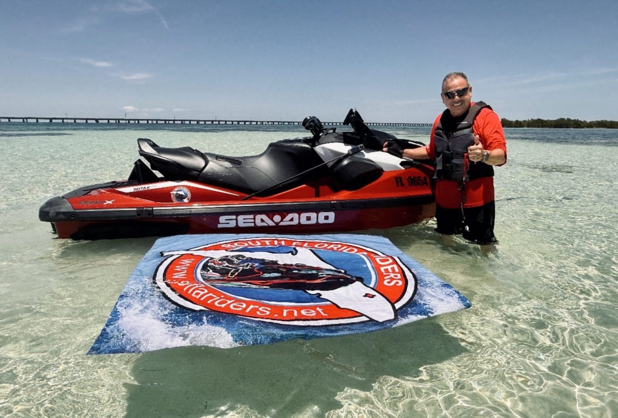 Screenshot - The Wake Edit Man in a red life vest stands in shallow tropical water next to a red Sea-Doo jet ski, giving a thumbs up with a long bridge in the background.