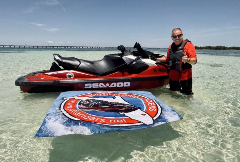 Man in a red life vest stands in shallow tropical water next to a red Sea-Doo jet ski, giving a thumbs up with a long bridge in the background.