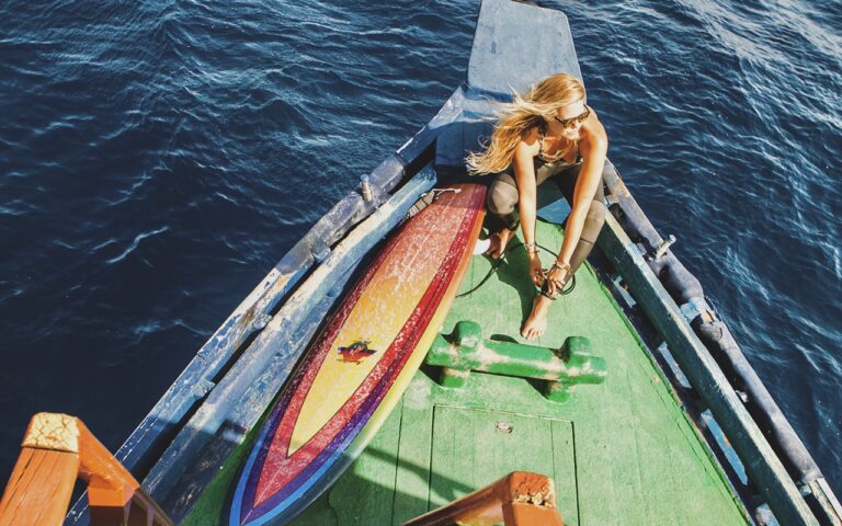 Woman wearing sunglasses sits on the bow of a small boat with a rainbow-colored surfboard beside her as the blue ocean surrounds them.