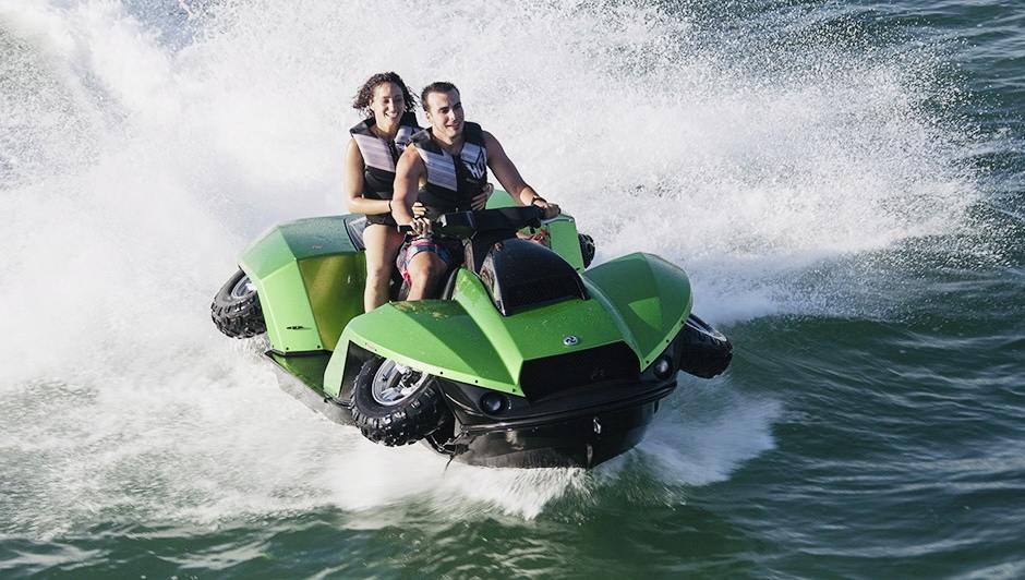 MAIN - The Wake Edit Two people wearing life jackets ride a bright green jet ski as it jumps over a wave, spraying water around them.
