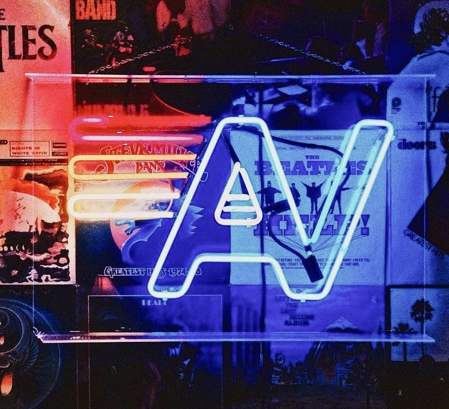 Unknown - The Wake Edit Blue neon sign in the shape of the letter A against a collage of posters in a dark room.