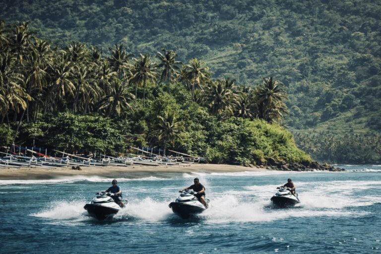 Three people riding jet skis on blue water near a tropical beach with palm trees and green hills in the background.
