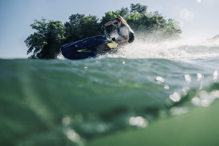 Person wearing sunglasses on a blue WaveRunner jet ski, speeding across a lake with trees in the background.