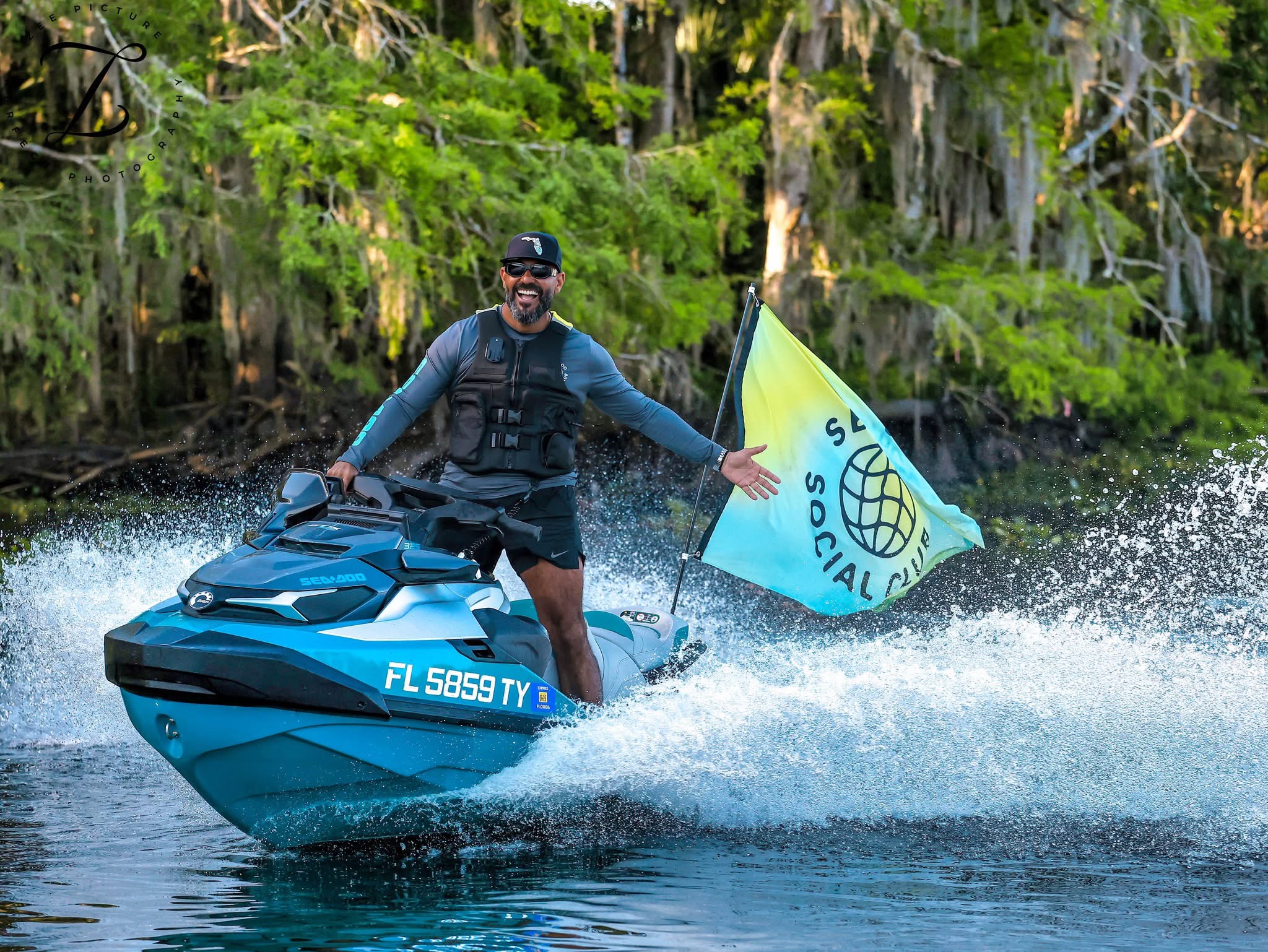 Man wearing life vest and sunglasses joyfully rides a blue jet ski through splashing water with a green forest backdrop and a turquoise flag nearby