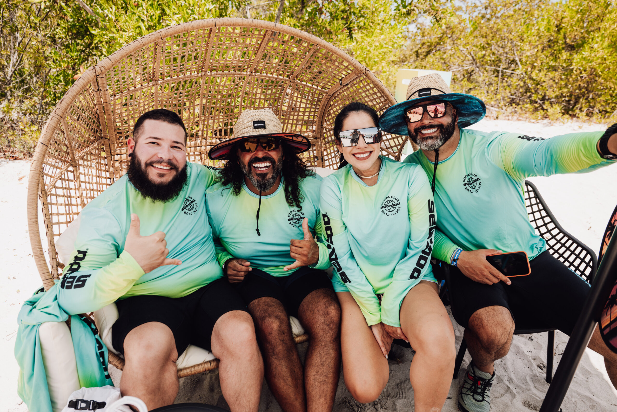 Sea-Doo Legendary Meetup - The Wake Edit Four friends wearing matching teal shirts sit close together in a large wicker beach chair, smiling for a selfie on the sunny sand.