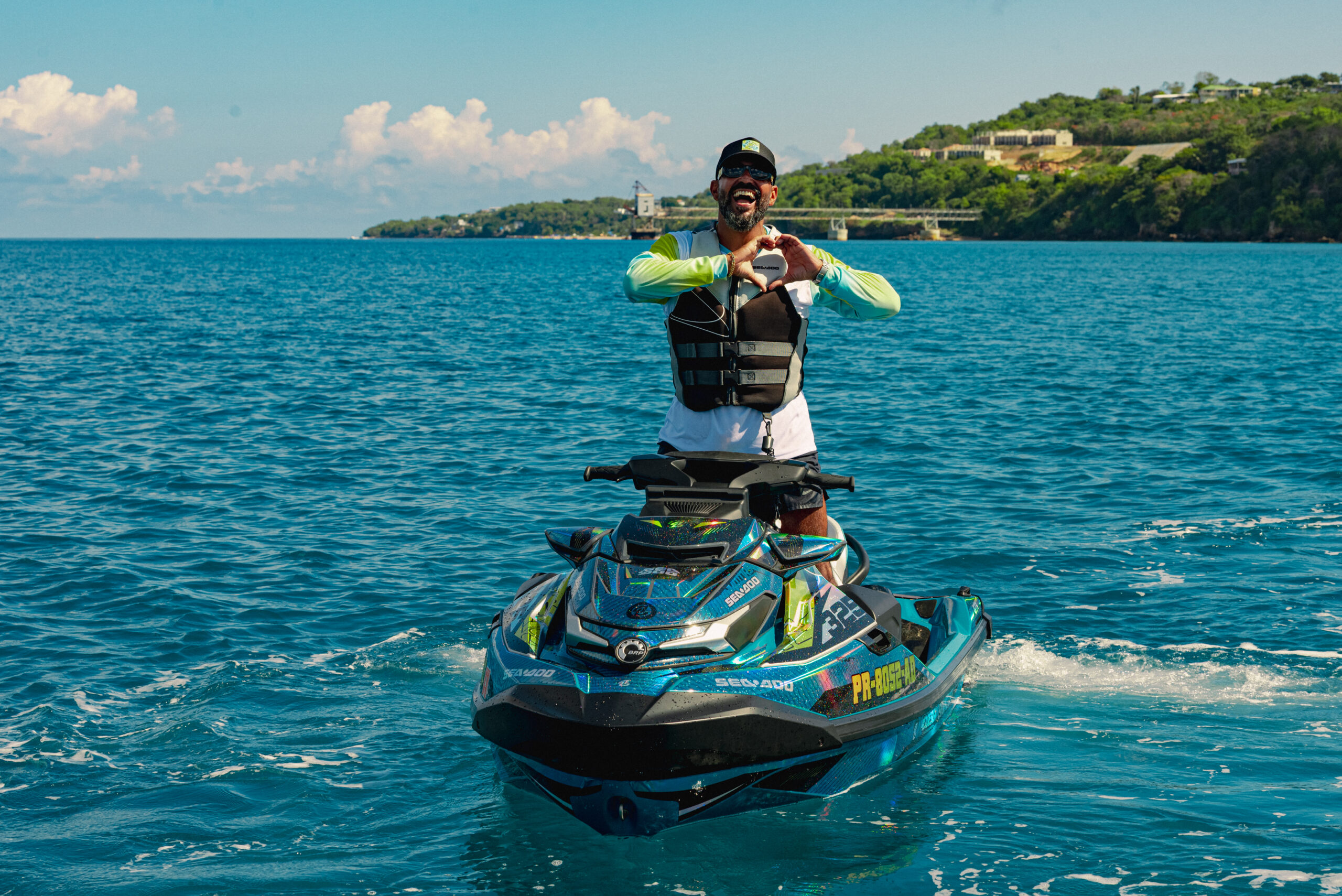 Man on a jet ski wearing a life jacket, forming a heart shape with his hands on blue ocean with a distant coastline.