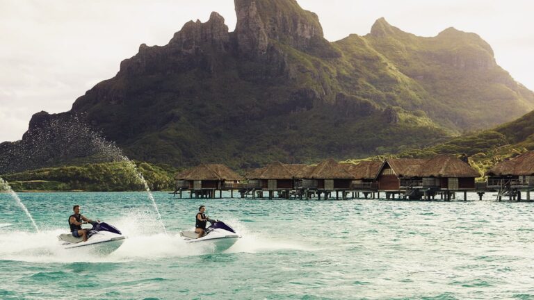 Two people ride white and purple jet skis across turquoise water near overwater bungalows with a lush mountain backdrop.