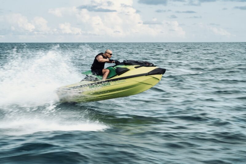 MAIN - The Wake Edit A bald man wearing a life jacket rides a yellow-green jet ski at high speed across the blue ocean, spraying water behind him.