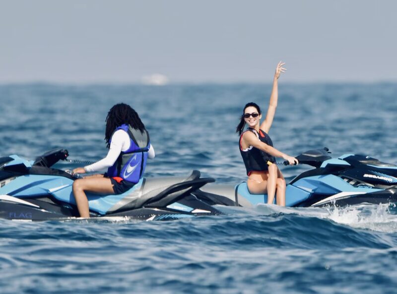 Main - The Wake Edit Two people ride blue jet skis on the ocean; the woman on the right waves and smiles at the camera.