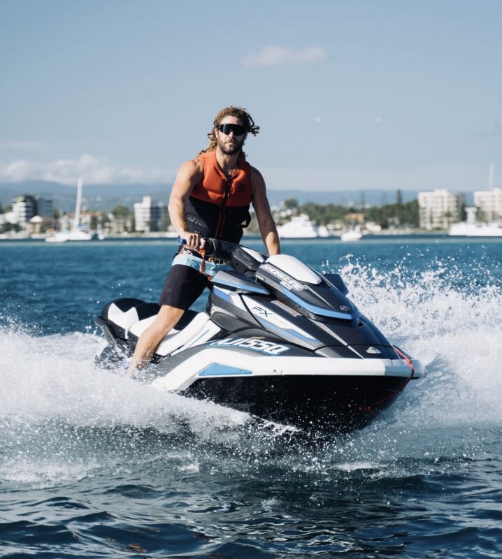 Man wearing an orange life vest rides a white and blue jet ski across blue water with a city skyline in the background.
