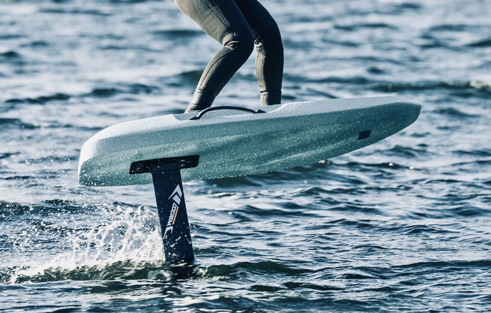 IMAGE 2 - The Wake Edit Person riding a hydrofoil board above the water, skimming with a foil beneath the board. In a wetsuit on a sunny day at sea.