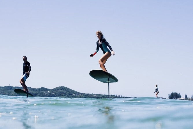 IMAGE 3 - The Wake Edit Woman on a hydrofoil board gliding above the turquoise water, others surfing in the background on a sunny day beach.