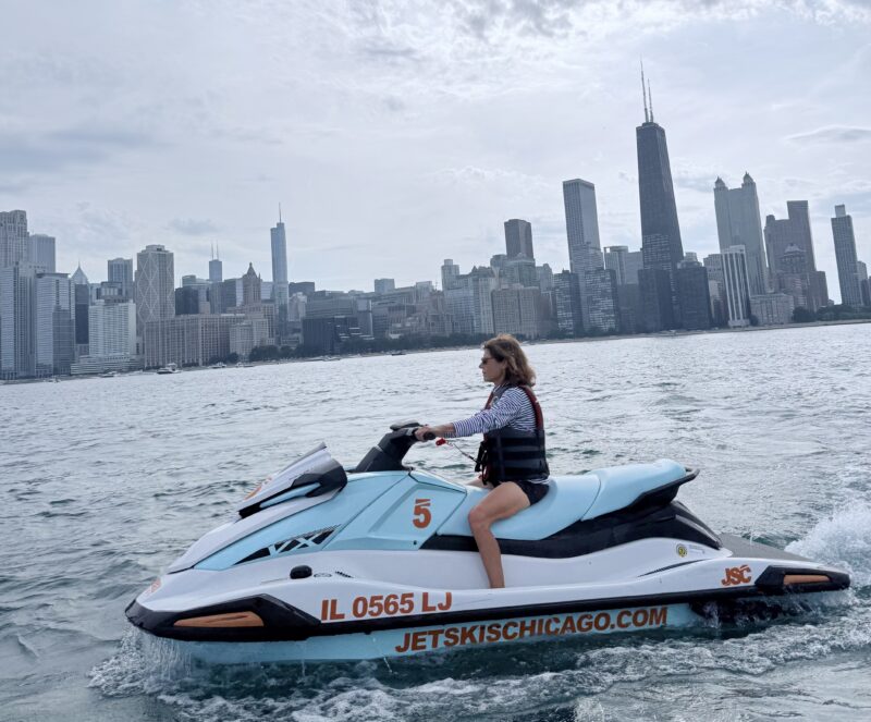 Person rides a blue and white jet ski on the water with a city skyline in the background.