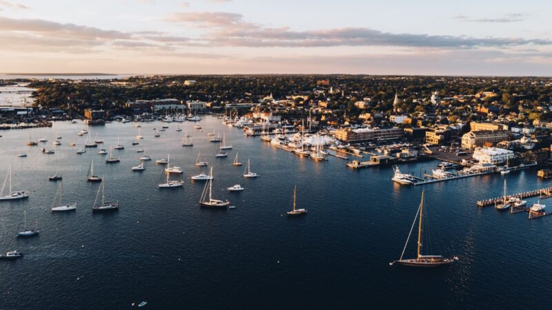 MAIN - The Wake Edit Aerial view of a harbor filled with numerous sailboats and a seaside town at sunset. (Informative; concise)