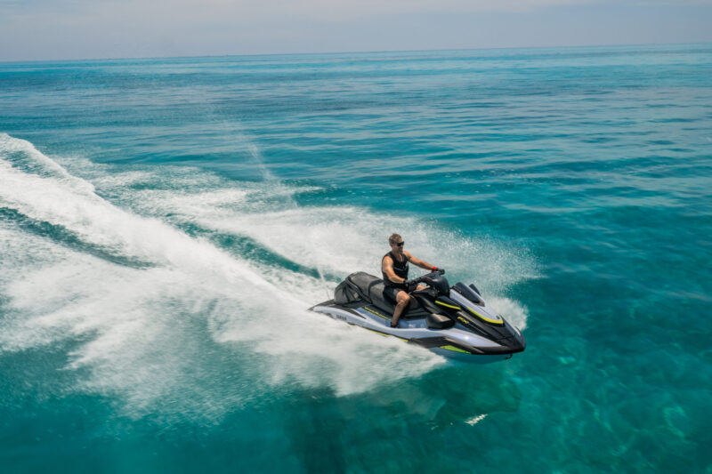 Man riding a black and white jet ski across turquoise ocean, leaving a white wake behind him.